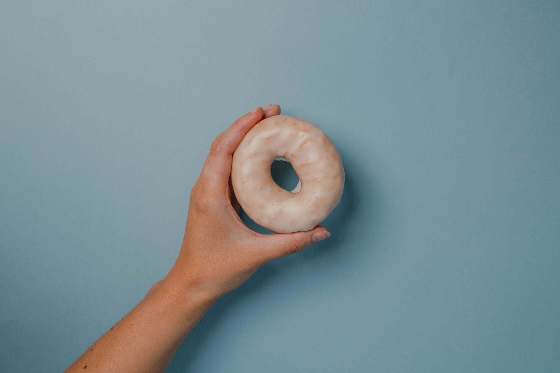 A hand holding a glazed gluten-free Happy Dough doughnut against a blue backdrop